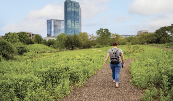 A woman walks a bark-covered path in an urban greenbelt, with tall buildings in the background