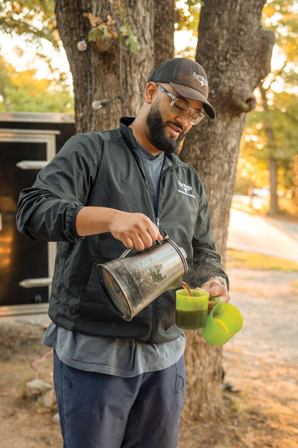 Man in a hat pouring coffee into one mug while holding another at a campsite