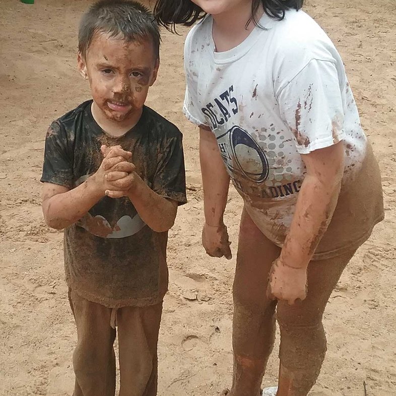 Girl in white playing in the mud with younger sibling who is dressed in black.